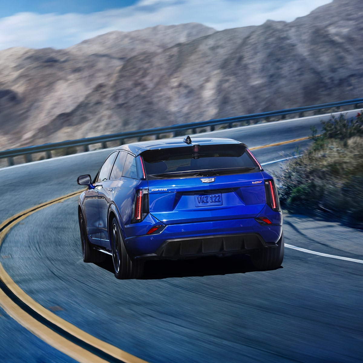 Rear View of a Blue 2026 OPTIQ-V Driving on a Curve of a Mountain Row